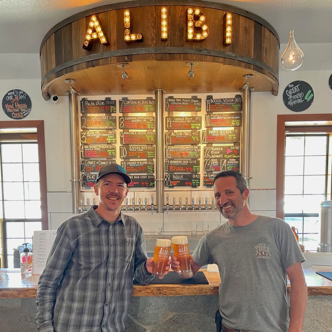Alibi Ale Works founders holding beers beneath the Alibi sign and tap list